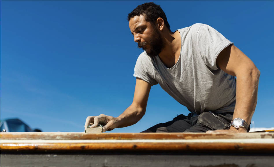 A young man sands a wooden boat deck in the hot sun